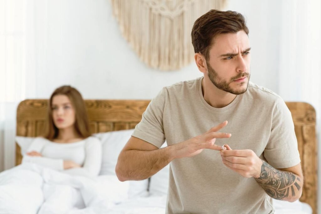 Uomo con una fede nuziale in mano mentre riflette sulla fine della relazione, con la compagna sfocata sullo sfondo.